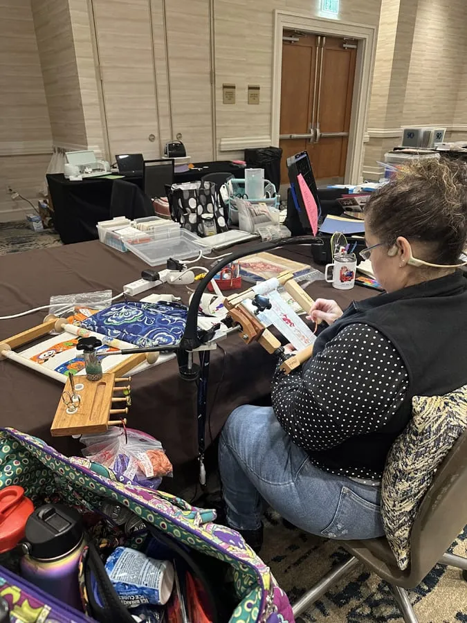 A retreat-goer at her table working a cross-stitch piece on a scroll frame, with a thread organizer and a Cricut on the tables behind her