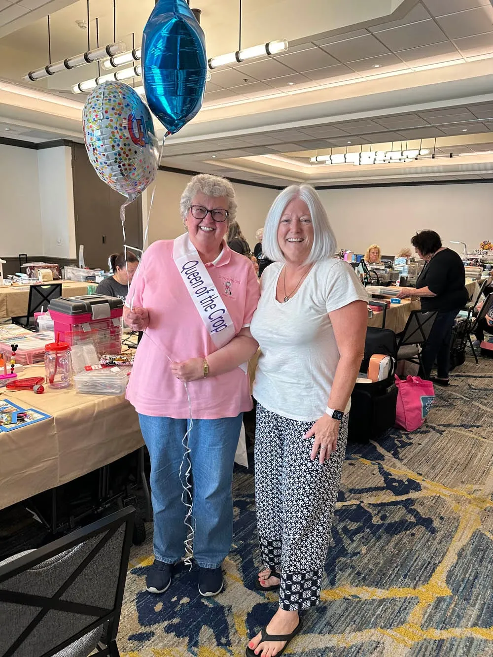 A Queen of the Crop winner in a pink polo and sash, balloons in hand, posing with a friend in the crop room