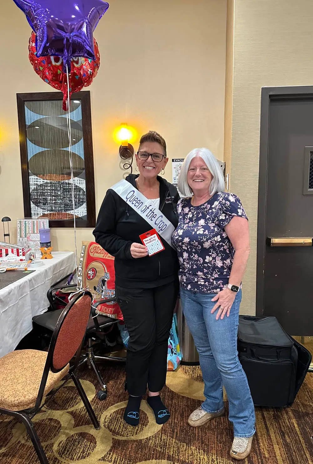 The Queen of the Crop in her sash, holding her prize beside a friend under celebration balloons