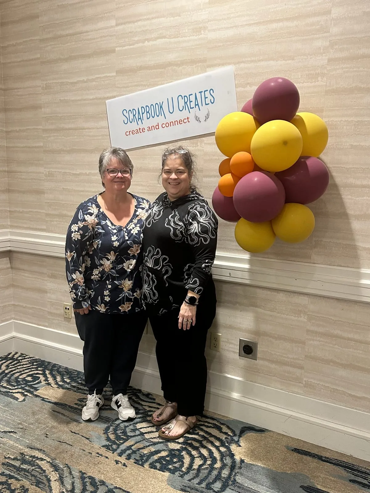 Two friends smiling beside the Scrapbook U Creates welcome sign and a cluster of yellow and plum balloons