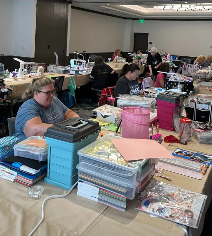 A crop retreat in full swing: rows of six-foot tables in a hotel ballroom, and a smiling attendee in the foreground surrounded by paper, storage cases, and supplies