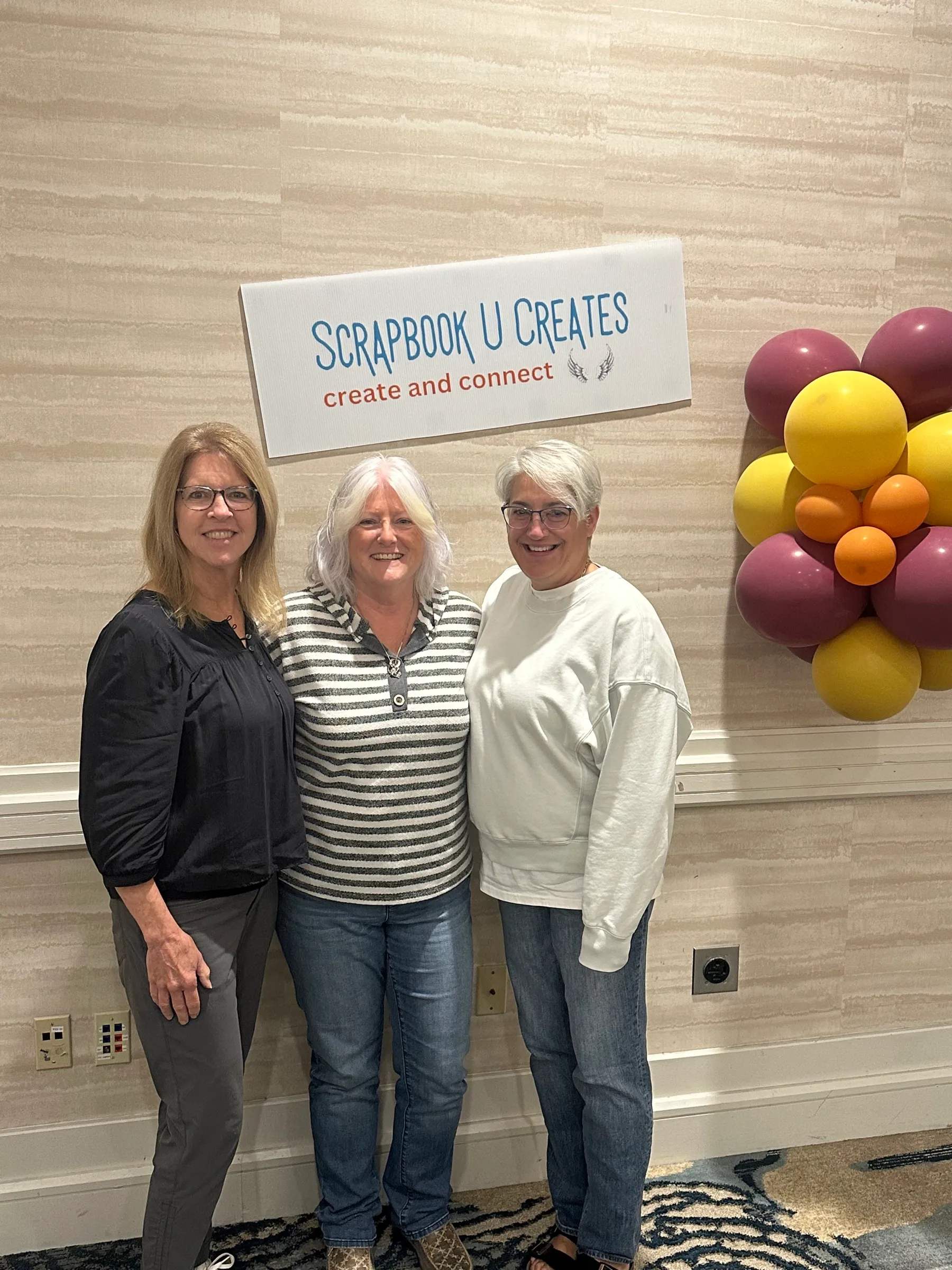 Three smiling retreat-goers standing in front of a Scrapbook U Creates sign and a cluster of plum, yellow, and orange balloons